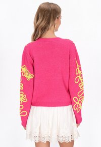 Rožinė megztinė ilgomis rankovėmis, papuošta geltonomis dekoratyvinėmis spiralėmis ant rankovių; tekstūruoto mezgimo audinys, apvalus kaklo iškirpimas ir raukšlėta apdaila.