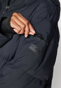 Black puffer jacket features a zippered pocket on the left sleeve and a textured Roxy logo patch, showcasing a sleek design and material.