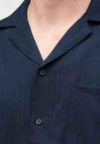 Πουκάμισο σε σκούρο μπλε χρώμα με κουμπιά, με γιακαδάκι και ορατή τσέπη στο στήθος, φορεμένο από άτομο με ανοιχτόχρωμο δέρμα.