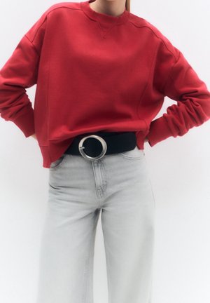 Personne portant une chemise rouge à manches longues rentrée dans un jean gris clair avec une ceinture noire avec une grande boucle circulaire argentée.