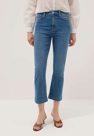 Donna che indossa jeans blu a vita alta e alla caviglia, camicetta bianca a maniche lunghe e sandali con tacco marroni e punta aperta, in piedi su uno sfondo semplice.