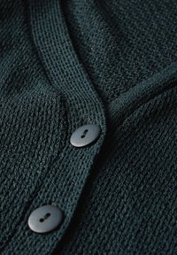 Stickad mörk teal cardigan med ett texturerat mönster, som har tre runda svarta knappar längs framknäppningen.