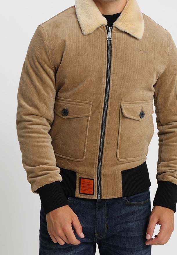 Homme portant une veste en velours côtelé beige avec col en peau lainée, fermeture éclair à l'avant, poches à boutons, poignets noirs côtelés et jean bleu.