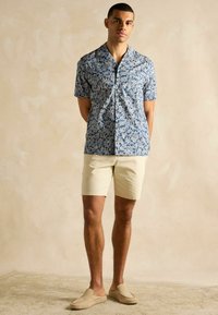 Košeľa s krátkym rukávom v modrej farbe s bielou kvetinovou potlačou, krémové šortky a svetlo béžové slip-on topánky; model stojí s rukami zloženými za chrbtom.