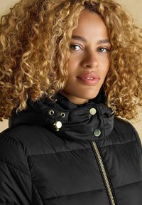 Kaskjutuudega blond naine, kes kannab musta puhujakihti kuldse tõmbluku ja nööpidega, beežil taustal.