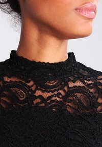 Sort blonde tøj med høj hals, intrikat blomstermønster og struktureret stof, der afslører en glat hudtone nedenfor.