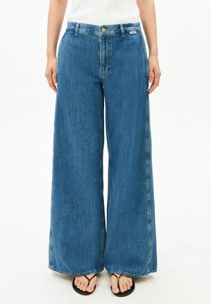 Vrouw draagt hoge taille, wijd uitlopende blauwe jeans gecombineerd met zwarte thongsandalen tegen een witte achtergrond.