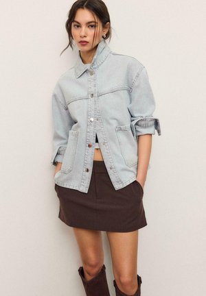 Mladi žena nosi svijetlu traper jaknu s izvrnutim rukavima, smeđu suknju sa uskim prugama i smeđe čizme, stoji nasuprot jednoličnoj pozadini.