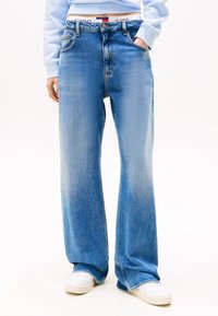 Světle modré široké džínsy vyrobené z denimu, obsahující klasický design s pěti kapsami a páskem se značkovým logem, kombinované s bílými teniskami.
