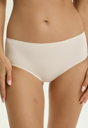 Kobieta w bezszwowej białej bieliźnie typu hipster, ukazująca brzuch, pępek i górną część ud, z zadbanymi paznokciami opartymi na biodrach.