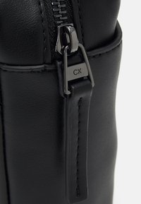 Primo piano di una borsa in pelle nera con una cerniera metallica e un'etichetta con il logo CK. La texture è liscia, con cuciture visibili e un design strutturato.