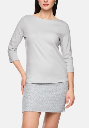Frau trägt ein hellgraues Oberteil mit Bootsausschnitt und Dreiviertelärmeln sowie einen strukturierten grauen Minirock vor einfarbigem Hintergrund.