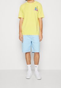 Camiseta de algodón amarilla con un pequeño estampado gráfico, combinada con pantalones cortos de color azul claro. El modelo lleva zapatillas blancas y calcetines de longitud media.
