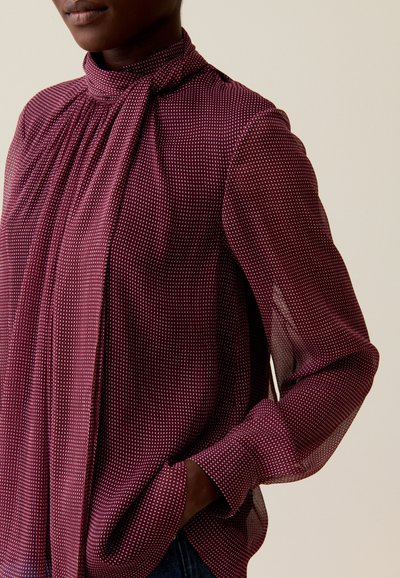 Mujer vistiendo una blusa larga de manga larga transparente en burdeos con un cuello alto y detalle de lazada, con una mano en el bolsillo, frente a un fondo liso.