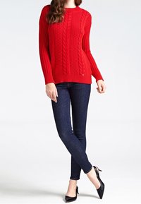Rode gebreide trui met een kabelontwerp, geribbelde textuur en lange mouwen, gecombineerd met donkere skinny jeans en zwarte puntige schoenen.