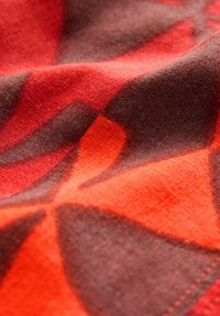 Textil har ett abstrakt mönster med orange och röda former på en brun bakgrund, som visar en mjuk textur och en slät yta.