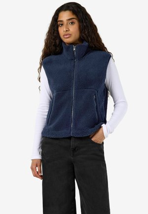Junge Frau mit langen lockigen Haaren, die eine marineblaue Fleecejacke mit Reißverschluss über einem weißen Langarmshirt und schwarzen Hosen trägt, steht mit einer Hand in der Tasche.