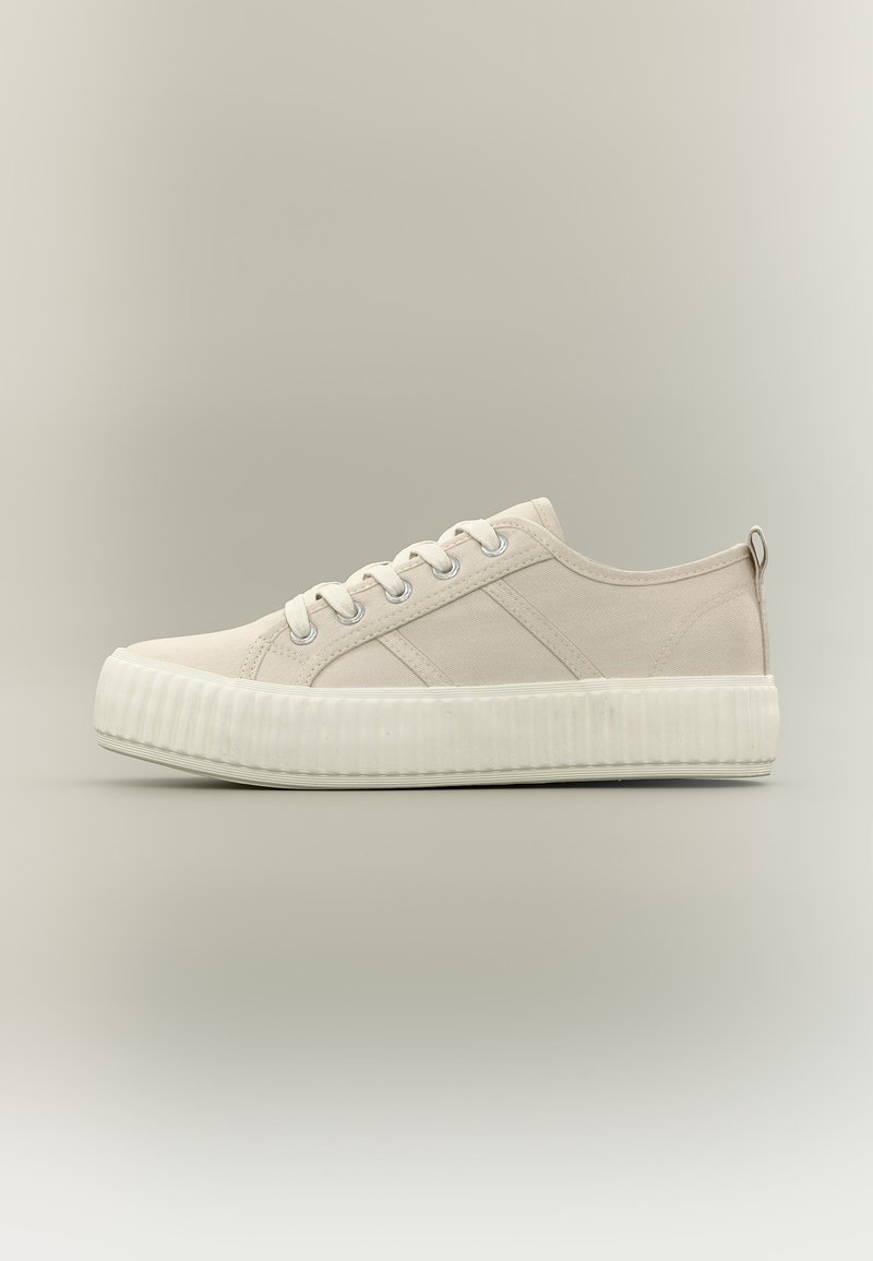 Μπεζ χαμηλού προφίλ καμβαδένιο αθλητικό παπούτσι με λευκή χοντρή ελαστική σόλα και κορδόνια, εμφανίζεται από το πλάι σε ουδέτερο φόντο.