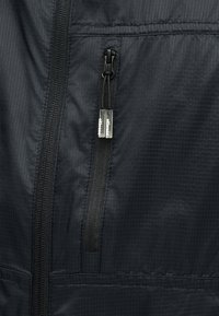 Gros plan sur une veste en tissu noir avec deux fermetures éclair verticales, une principale et une plus petite pour une poche, toutes deux équipées de tirettes argentées arborant la marque.