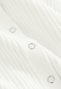 Weißes, geripptes Stoffmaterial mit silbernen Knopfakzenten. Nahaufnahme zeigt das strukturierte Muster und die Knopfdetails vor einer glatten Oberfläche.