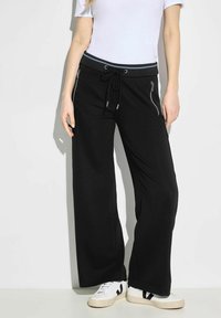 Svarta vida byxor i mjukt tyg, med dragsko i midjan och sidogylf. Matchas med vita sneakers.