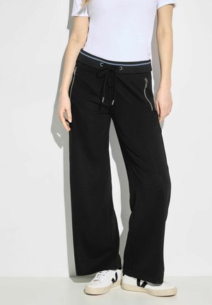Pantalones negros de pierna ancha hechos de tela suave, con una cinturilla elástica ajustable y cremalleras laterales. Combinados con zapatillas blancas.