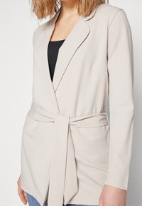 Beige wikkelblazer gemaakt van zachte stof, met een strikriem in de taille, lange mouwen en een borstzakje.