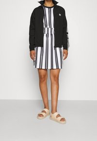 Svart zip-up jacka, vit- och svartrandig klänning med elastisk midja, och beige sandaler med utskärningar. Slätt tyg och avslappnad passform.