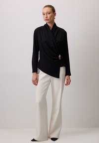 Melnas garās piedurknes blūze ar plūstošu dizainu un zelta aksesuāriem, apvienota ar augsto vidukli baltām biksēm un melniem stiletto papēžiem.