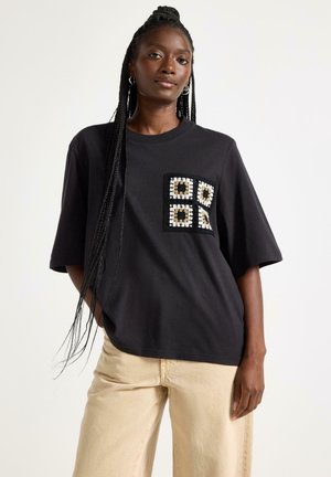 Kvinde med lange fletninger iført sort oversized T-shirt med hæklede firkantede lomme og beige bukser, stående mod en ensfarvet baggrund.