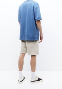 Hombre que lleva pantalones cortos beige claros, calcetines blancos, zapatillas en blanco y negro, y una camisa azul de manga corta holgada, de pie contra un fondo blanco.