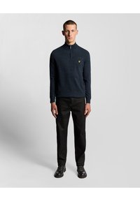 Mörkblå, långärmad zip-up-tröja med en texturerad yta och en liten gul logotyp på bröstet, kombinerad med svarta byxor och stövlar.