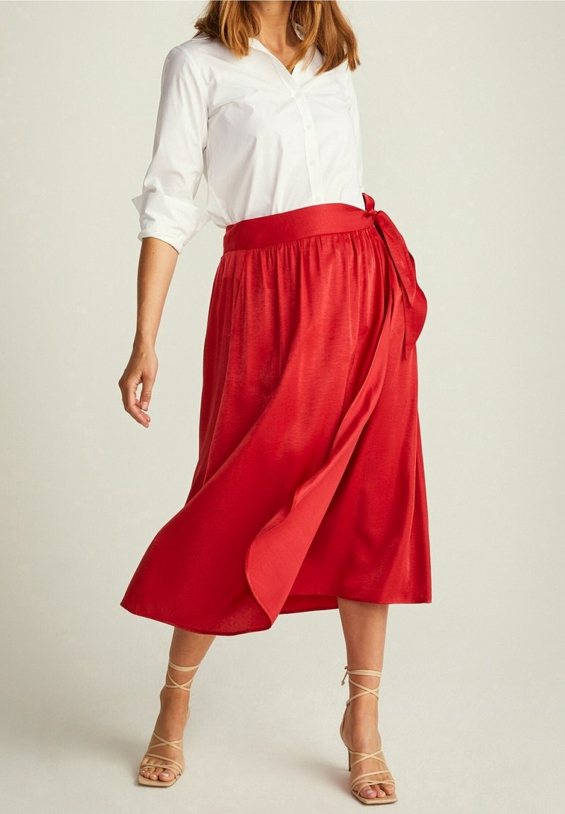 Falda midi roja y fluida con una amplia cinturilla y detalle de lazo en el frente, combinada con una camisa blanca de botones y tacones de tiras beige.