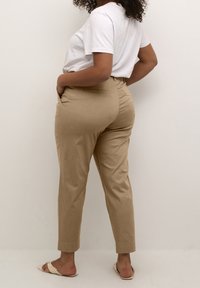 Beige skræddersyede bukser med en glat tekstur, sidelommer og en tætsiddende talje, parret med en hvid t-shirt og sandaler.