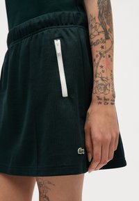 Falda de mezcla de algodón en verde oscuro con cinturilla elástica; cuenta con un bolsillo con cremallera blanca y un pequeño logotipo bordado cerca del dobladillo.