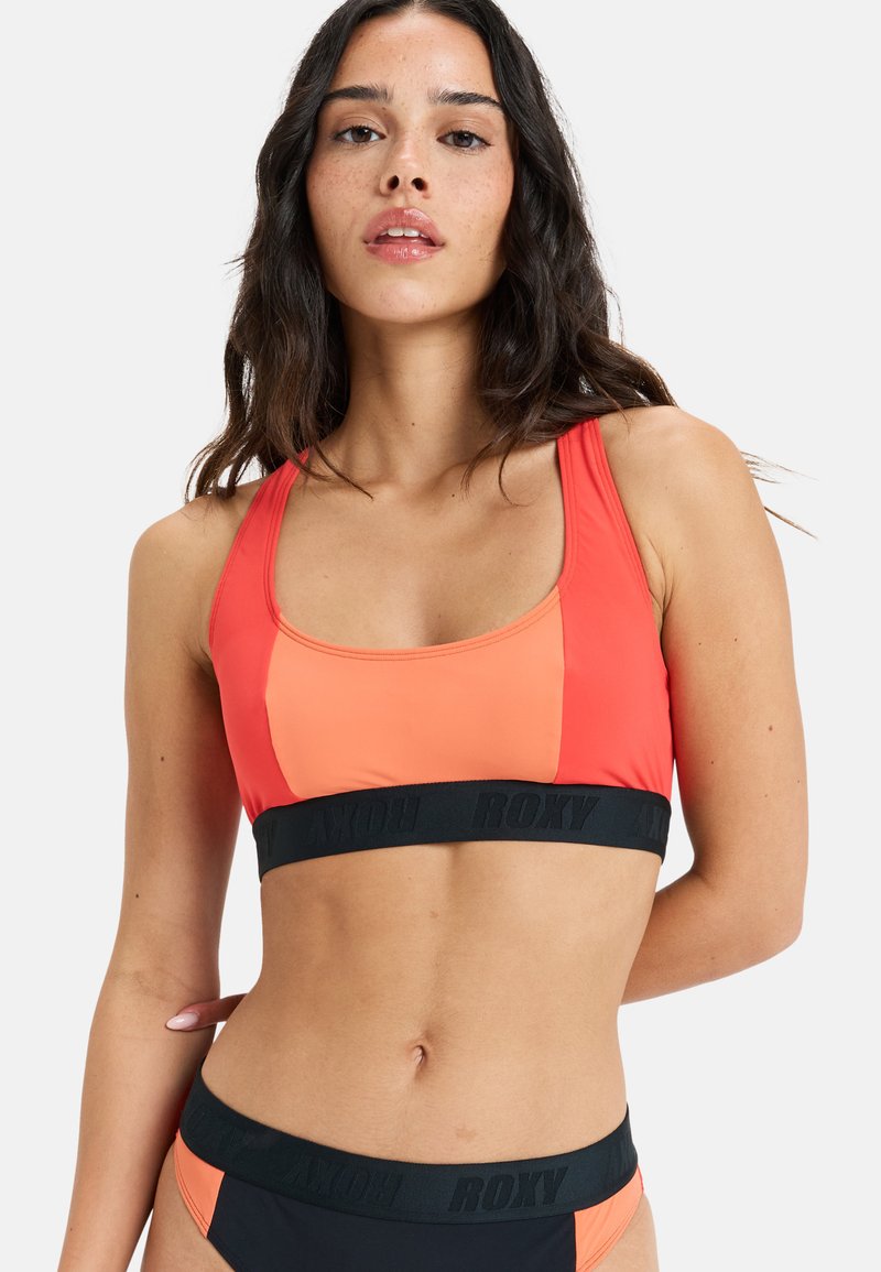 Sportlicher Bikini-Oberteil in Koralle und orangefarbenen Blöcken; mit einem schwarzen Band mit "ROXY"-Logo; der Stoff hat eine glatte, dehnbare Textur.