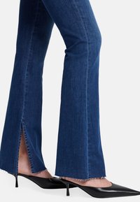 Niebieskie dżinsowe spodnie dzwony z bocznymi rozcięciami na końcu i postrzępionymi brzegami, w zestawie z czarnymi czółenkami na szpilce. Teksturowana tkanina z delikatnym połyskiem.