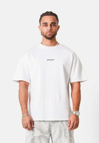 Hombre con cabello trenzado que lleva una camiseta blanca SIXTHJUNE, pantalones cortos gris claro con estampado, reloj plateado, anillo, pulsera, de pie contra un fondo liso.