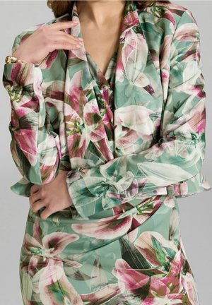 Frau, die einen pastellgrünen Blazer mit Blumenmuster und ein passendes Kleid mit pinken und weißen Lilien trägt, posiert mit verschränkten Armen und einer Hand nahe ihrem Hals.