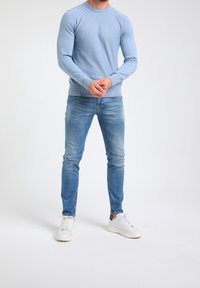 Mężczyzna w jasnoniebieskim, strukturalnym swetrze, dopasowanych, wyblakłych niebieskich dżinsach i białych trampkach, stoi z dłońmi złożonymi.