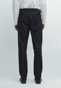Jeans denim scuri con una vestibilità slim, caratterizzati da una patta in pelle marrone in vita e due tasche posteriori. Indossati con una camicia bianca a maniche lunghe.