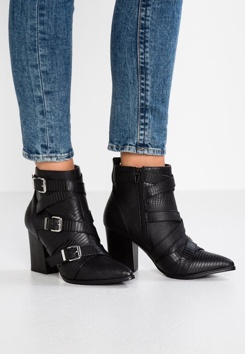 Bottes à chevilles texturées noires avec bouts pointus, talons épais, fermeture éclair latérale et trois sangles argentées à boucles, portées avec un jean en denim bleu.