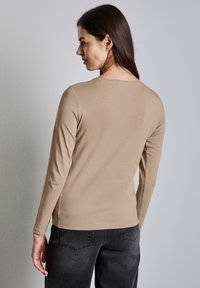 Langärmliges, hellbraunes Oberteil aus weichem Stoff, mit rundem Ausschnitt und figurbetonter Passform. Die Rückansicht zeigt eine glatte Textur und subtile Saumdetails.