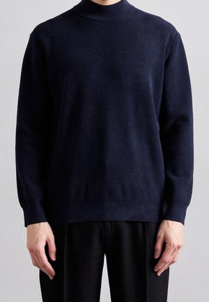 Personne portant un pull côtelé bleu marine foncé à manches longues avec un col montant et un pantalon noir, debout devant un fond gris clair.
