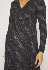 Svart långärmad klänning med V-ringning, som har en strukturerad tyg och metalliska guldprickar i ett abstrakt mönster. Empiremidja silhuett.