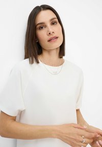 Hvid bluse med korte ærmer, rund halsudskæring og glat tekstur. Sølv lagdelte halskæder og ring fremhæver det samlede look.