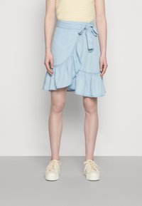 Saia envolvente de denim azul claro com bainha rendada, detalhe de laço na cintura e costura em contraste. Combinada com ténis de atacadores em tom creme.