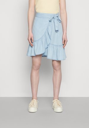 Saia envolvente de denim azul claro com bainha rendada, detalhe de laço na cintura e costura em contraste. Combinada com ténis de atacadores em tom creme.