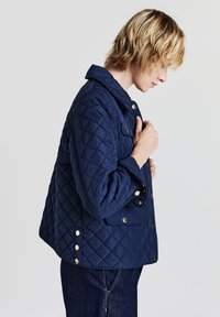 Una persona con capelli biondi corti che sistema il colletto di una giacca trapuntata blu navy con bottoni a pressione dorati, indossando jeans scuri.