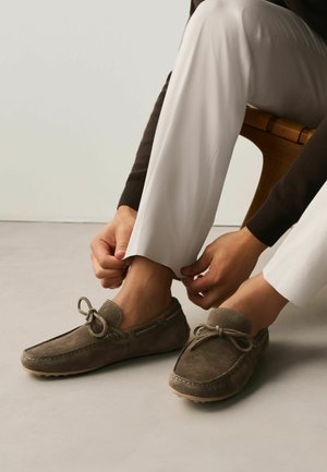 Person sitting on wooden chair adjusting white pants above brown suede loafers with bow details on light floor and plain background.